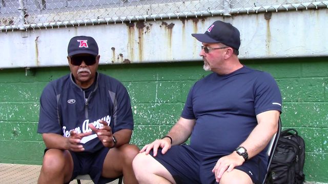 Reggie Smith, in a Team Avenue cap and jacket, talks with an interviewer in front of a green dugout wall.