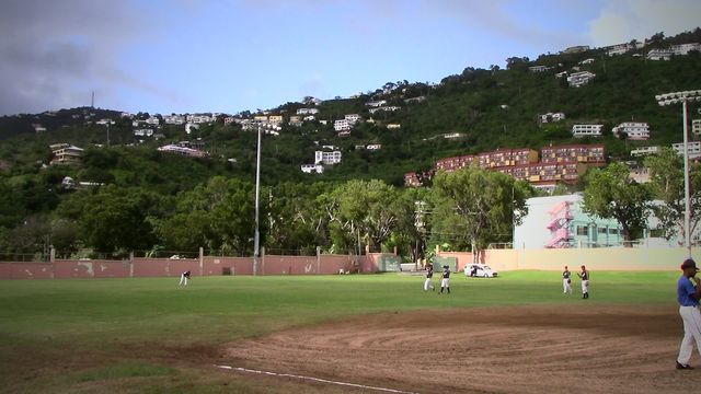 Wide shot of a baseball field below green hills and houses, with players spread across the infield.